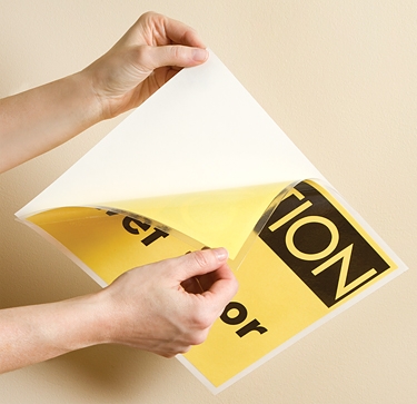 hands peeling backing sheet from laminated, adhesive-backed CAUTION sign
